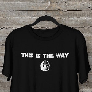 以下が含まれることがあります： 白い文字で「This is the way」と書いてある黒いTシャツと、白いマンダロリアンヘルメットのグラフィック