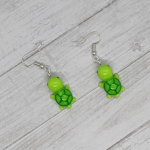 May include: A pair of green turtle dangle earrings with silver hooks. The turtles are made of polymer clay and have a glossy finish.