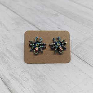 May include: A pair of iridescent blue and green spider earrings on a brown card.