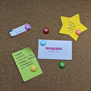 Op de afbeelding: Een kurkbord met verschillende items die eraan vastgeprikt zijn, waaronder een groene notitie met een boodschappenlijstje, een witte visitekaartje met de tekst "laceycakes make life sweeter", een gele stervormige notitie met de tekst "CALL YOUR MOM!" en verschillende kleurrijke M&M's.
