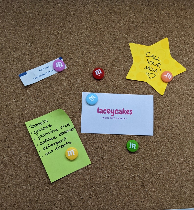 Op de afbeelding: Een kurkbord met verschillende items die eraan vastgeprikt zijn, waaronder een groene notitie met een boodschappenlijstje, een witte visitekaartje met de tekst "laceycakes make life sweeter", een gele stervormige notitie met de tekst "CALL YOUR MOM!" en verschillende kleurrijke M&M's.