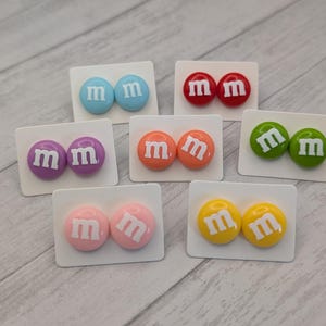 May include: A collection of colorful stud earrings, each featuring a pair of round, candy-like designs. The earrings are in various colors, including blue, red, purple, orange, green, pink, and yellow, each with a white "m" on them.