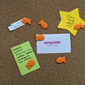 May include: A corkboard with various items pinned to it, including a green note with a list of items, a white business card with the text "laceycakes make life sweeter", a yellow star-shaped note with the text "CALL YOUR MOM!" and a heart, and several orange fish-shaped crackers.