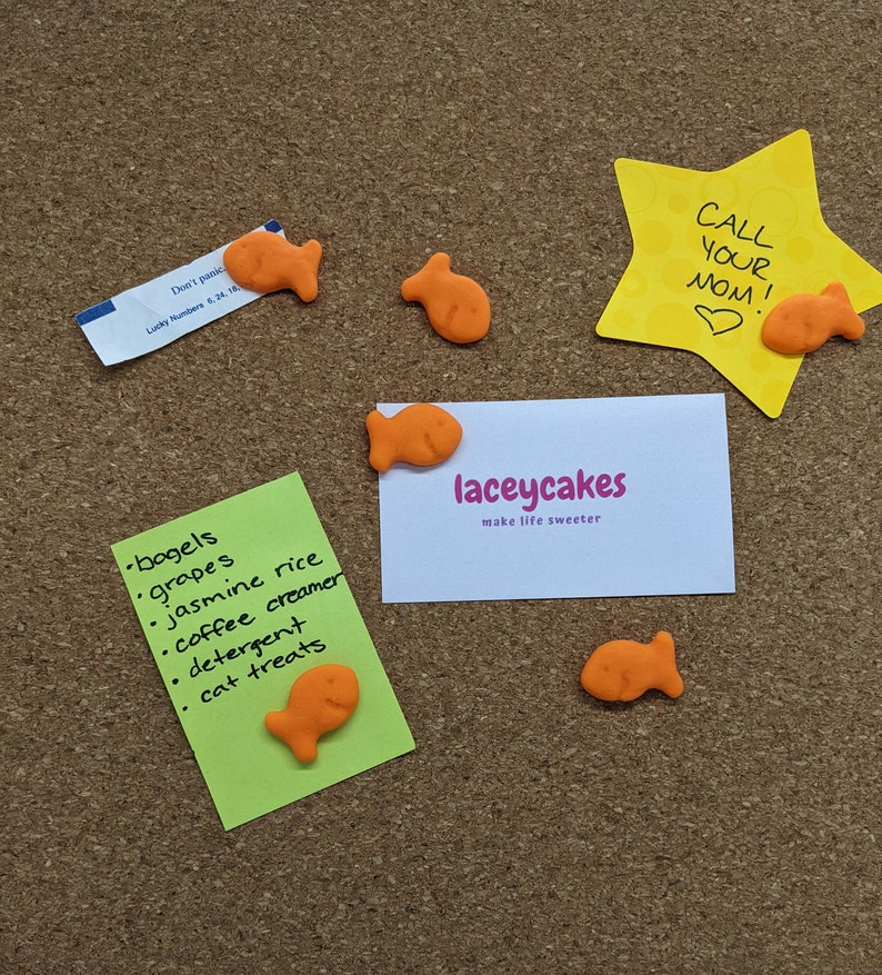 May include: A corkboard with various items pinned to it, including a green note with a list of items, a white business card with the text "laceycakes make life sweeter", a yellow star-shaped note with the text "CALL YOUR MOM!" and a heart, and several orange fish-shaped crackers.