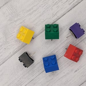 May include: Six colorful plastic push pins shaped like Lego bricks. The push pins are yellow, green, purple, red, blue, and black.