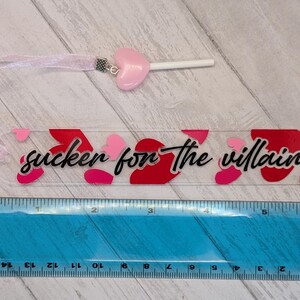 May include: A clear acrylic bookmark with the text "sucker for the villain" in black script, decorated with red and pink hearts. A pink heart-shaped charm and ribbon are attached. The bookmark is approximately 6 inches long.
