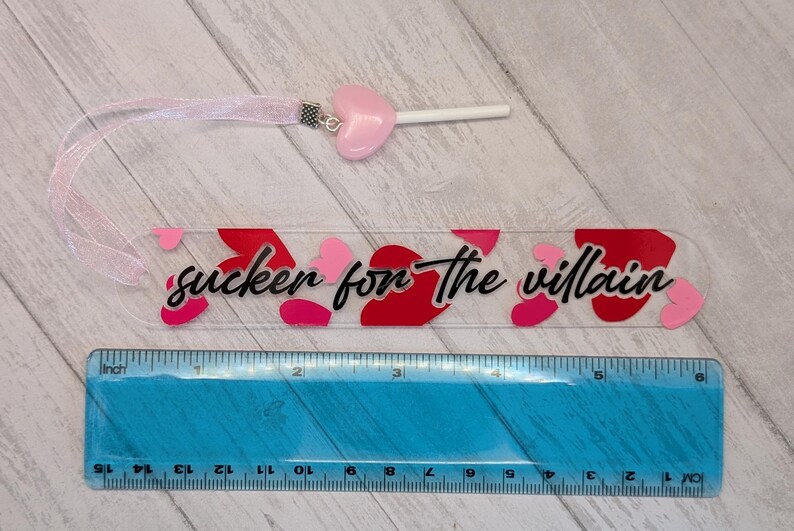 May include: A clear acrylic bookmark with the text "sucker for the villain" in black script, decorated with red and pink hearts. A pink heart-shaped charm and ribbon are attached. The bookmark is approximately 6 inches long.