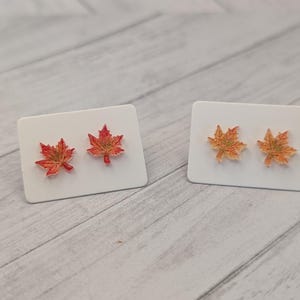 May include: Two pairs of maple leaf stud earrings. One pair features red and orange leaves, the other pair has orange leaves. The earrings are displayed on white rectangular cards, perfect for fall.