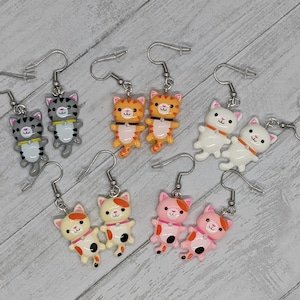 May include: A set of 10 cat-shaped earrings. The earrings are made of polymer clay and come in a variety of colors, including gray, orange, white, and pink. Each cat is wearing a collar with a small bell.