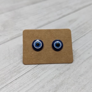 May include: A pair of blue evil eye stud earrings on a brown card. The earrings are round and have a white center with a black pupil.
