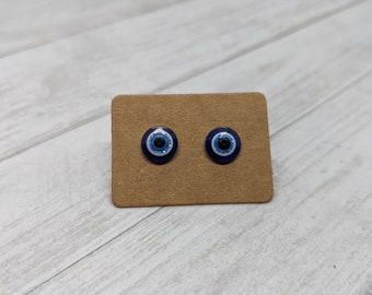 May include: A pair of blue evil eye stud earrings on a brown card. The earrings are round and have a white center with a black pupil.