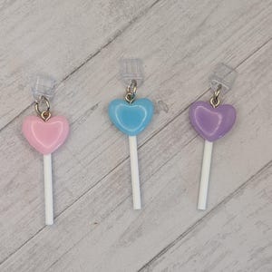 May include: Three heart-shaped lollipop earrings in pink, blue, and purple. Each lollipop has a white stick and a silver-colored earring hook. The earrings are displayed on a light wood-grain surface.