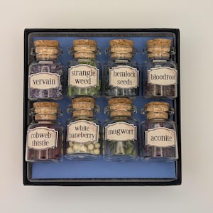 May include: A set of eight small glass bottles with cork stoppers, each labeled with a different herb name. The bottles are arranged in a black box with a blue interior. The herbs include vervain, strangle weed, hemlock seeds, and bloodroot.