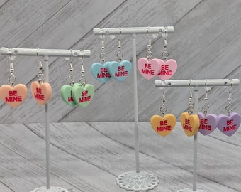 May include: A set of 10 heart-shaped earrings with the words "Be Mine" printed on them. The earrings are in various colours, including pink, blue, green, yellow, and purple.