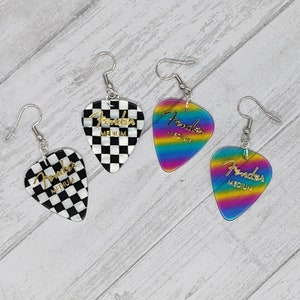 May include: Three pairs of guitar pick earrings. One pair is black and white checkered with gold lettering that says "Fender Medium". Another pair is rainbow with gold lettering that says "Fender Medium". The last pair is also rainbow with gold lettering that says "Fender Medium".