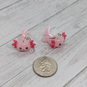 以下が含まれることがあります： ピンクのアホロートルイヤリングのペア。各イヤリングは、濃いピンクのエラと白い頭のクレストを持つ、小さな笑顔のアホロートルが特徴です。イヤリングは光沢のある素材でできており、シルバーカラーのイヤーフックが付いています。