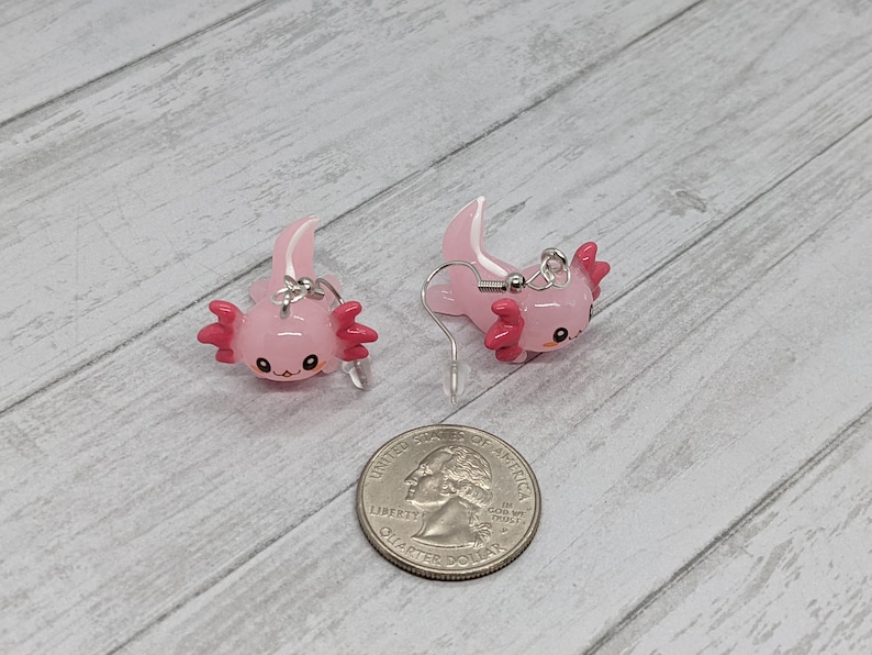 以下が含まれることがあります： ピンクのアホロートルイヤリングのペア。各イヤリングは、濃いピンクのエラと白い頭のクレストを持つ、小さな笑顔のアホロートルが特徴です。イヤリングは光沢のある素材でできており、シルバーカラーのイヤーフックが付いています。