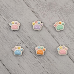 May include: Six pastel-colored paw print embellishments. Each paw print has a white base with a different color combination of pink, blue, yellow, orange, and green pads. The embellishments are arranged on a light gray wooden surface.