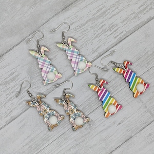 May include: A set of six bunny-shaped earrings with different patterns. Two earrings feature a plaid pattern in pink, blue, and green. Two earrings feature a floral pattern in pink, yellow, and green. Two earrings feature a rainbow stripe pattern. Each earring has a white pom-pom on the back.