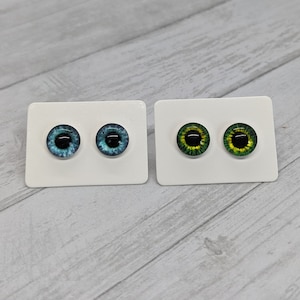 May include: Two pairs of glass eyes on white cards. One pair is blue with black pupils, the other is green with black pupils. The eyes are round and have a reflective quality, suitable for crafts or doll making.