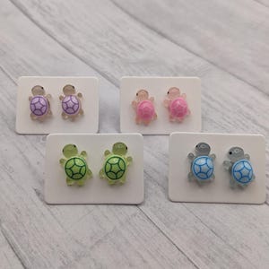 May include: Four pairs of stud earrings featuring a turtle design. The turtles are made of plastic and come in four colors: purple, pink, green, and blue.