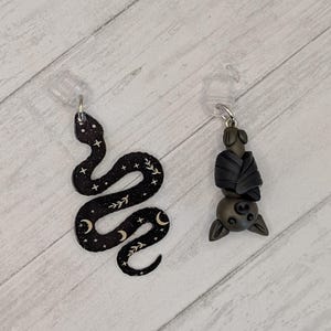 May include: A black snake charm featuring white celestial patterns of stars, moons, and leaves, paired with a small, three-dimensional dark gray bat charm. Both charms are attached to clear plastic clips for hanging or attachment purposes.