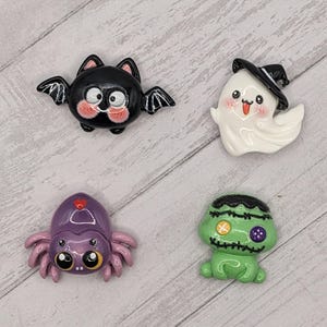 May include: Four Halloween-themed figurines: a black bat with pink cheeks, a white ghost wearing a witch hat, a purple spider, and a green Frankenstein's monster. Each figurine has a cartoonish design and is set against a light wood grain background.