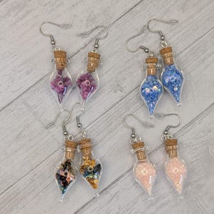 May include: A collection of six pairs of handmade earrings. Each earring features a small, clear glass bottle with a cork stopper. The bottles are filled with various colorful elements, including dried flowers, glitter, and small beads. The earrings have silver-colored ear wires.