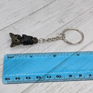 May include: A brown and black bat-shaped keychain with a silver chain and ring. The keychain is about 1 inch long and is measured against a ruler.