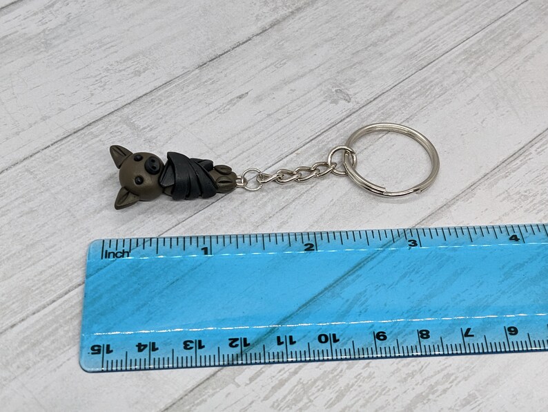 May include: A brown and black bat-shaped keychain with a silver chain and ring. The keychain is about 1 inch long and is measured against a ruler.