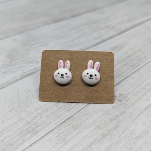 May include: A pair of white bunny stud earrings with pink ears and black eyes. The earrings are on a brown card.