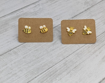 Può includere: Due paia di orecchini a bottone a forma di ape su cartoncino marrone. Un paio presenta un design di ape semplice, mentre l'altro paio presenta un'ape con le ali.