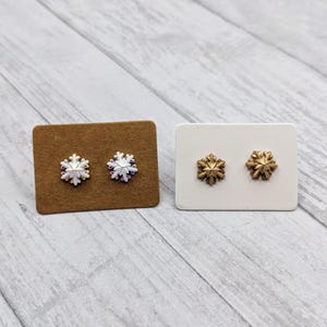 May include: Two pairs of snowflake stud earrings. One pair is silver with a white snowflake design. The other pair is gold with a gold snowflake design.