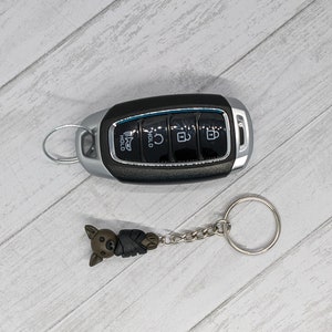May include: A black car key with four buttons and a silver ring. A brown and black cartoon animal keychain is attached to the key ring.