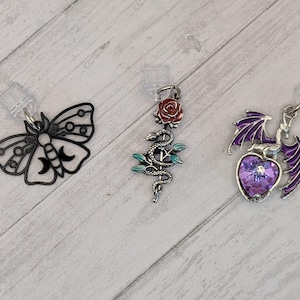 May include: Three decorative charms with clear plastic attachments. One is a black moth, another features a snake and red rose, and the third has a purple heart with a dragon. The charms are silver-toned with colorful enamel accents.