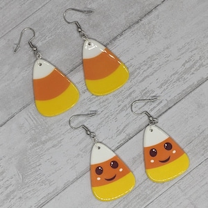 May include: A pair of dangle earrings with a candy corn design. The candy corn is white, orange, and yellow. One pair has a smiling face.