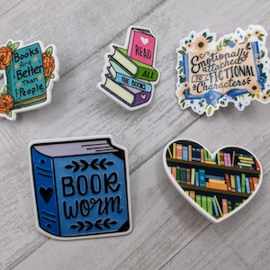 May include: Five colorful book-themed enamel pins. The pins feature various book-related quotes and illustrations, including a stack of books with the text "Read All The Books", a heart-shaped bookshelf, and a book with the text "Book Worm".