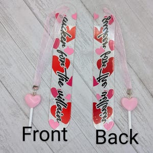 May include: Two clear acrylic bookmarks with pink ribbons and heart charms. The front bookmark has the text "I'm a sucker for the villain" with red and pink hearts. The back bookmark has the same text. The bookmarks are on a light wood surface.