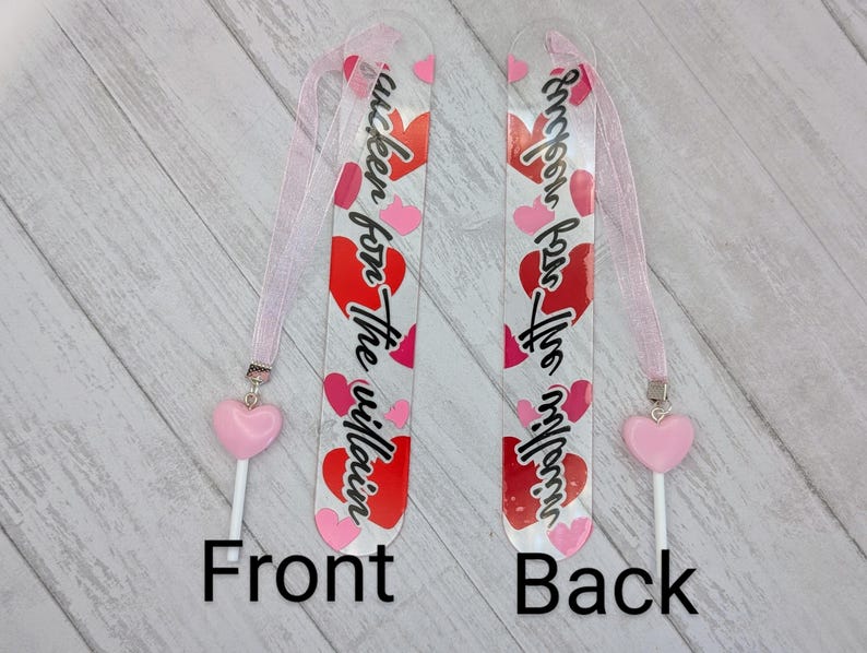May include: Two clear acrylic bookmarks with pink ribbons and heart charms. The front bookmark has the text "I'm a sucker for the villain" with red and pink hearts. The back bookmark has the same text. The bookmarks are on a light wood surface.