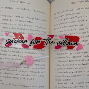 May include: A clear acrylic bookmark with the text "sucker for the villain" in black script, accented with red and pink heart shapes. A pink heart charm hangs from the bottom. The bookmark is placed in an open book.