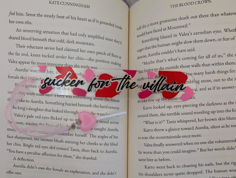 May include: A clear acrylic bookmark with the text "sucker for the villain" in black script, accented with red and pink heart shapes. A pink heart charm hangs from the bottom. The bookmark is placed in an open book.