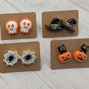 May include: Four pairs of Halloween-themed stud earrings on brown cards. Designs include white skulls with orange accents, black witch hats, white spiderwebs with black spiders, and orange jack-o'-lanterns with black cats.