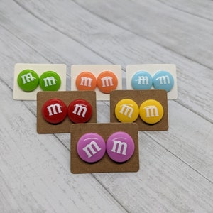 May include: A set of 10 colorful stud earrings featuring a white "M" on each earring. The earrings are displayed on brown card stock.