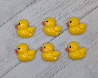 May include: Six yellow rubber duck decorations with orange beaks and black eyes are arranged on a light gray and white wooden surface. The ducks are facing different directions.