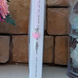 May include: A white book titled "THE BLOOD ORCS" with a pink ribbon bookmark and a heart-shaped charm. The author's name, Kate Cunningham, is printed at the bottom. The bookmark is attached to the top of the book.
