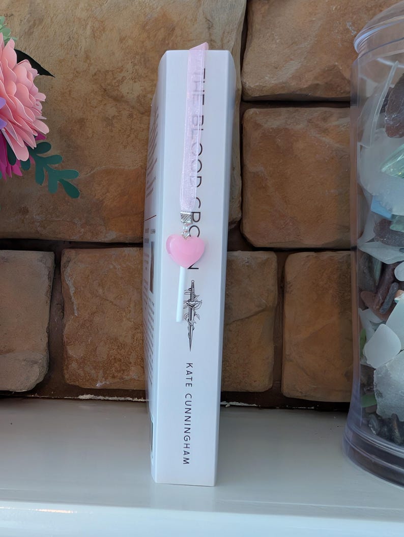 May include: A white book titled "THE BLOOD ORCS" with a pink ribbon bookmark and a heart-shaped charm. The author's name, Kate Cunningham, is printed at the bottom. The bookmark is attached to the top of the book.