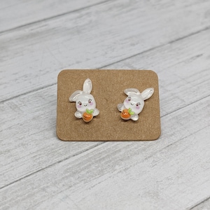May include: A pair of white bunny rabbit stud earrings with a small orange carrot in each bunny's hand. The earrings are on a brown card.