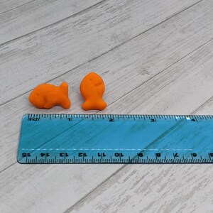 May include: Two orange fish-shaped crackers on a blue ruler with inch markings. The ruler is on a light wood surface.