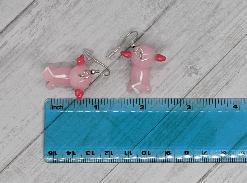 以下が含まれることがあります： ピンクのアホロートルイヤリングのペア。各イヤリングは、赤いエラと白いアクセントが付いた、小さく半透明のピンクのアホロートルが特徴です。イヤリングは、銀色のフィッシュフックのイヤワイヤーから吊り下げられています。イヤリングの長さは約2.5cmです。