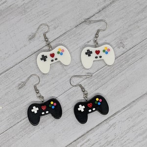 May include: A pair of black and white video game controller earrings. The earrings are silver and have a hook closure. The controllers have a red heart button and colorful buttons.